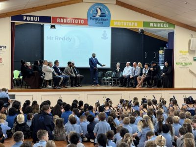 Man speaking on stage with rows of people to the left and right, lots of children looking at him from the audience