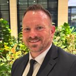 man in suit smiling in front of garden with building in background